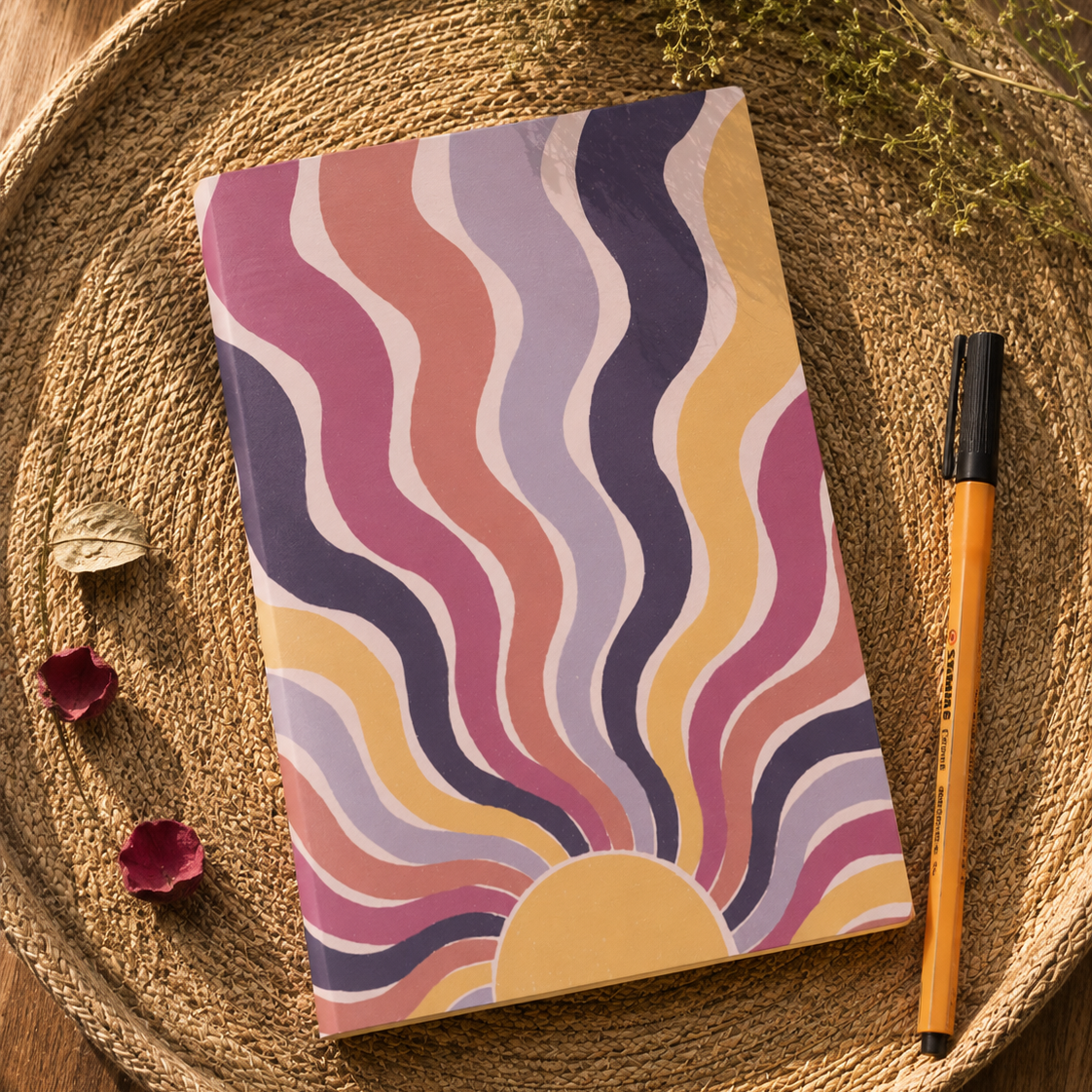 A notebook with a retro wavy sunburst pattern in pink, purple, yellow, and beige, styled on a woven round mat with a pen and dried botanicals.