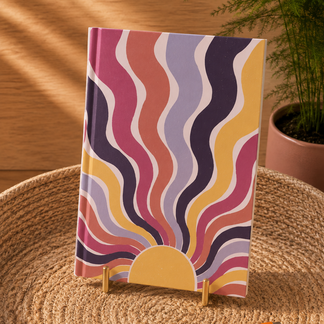 A notebook with a retro wavy sunburst pattern in pink, purple, yellow, and orange, displayed on a small wooden stand on a woven placemat with an orange pen nearby.